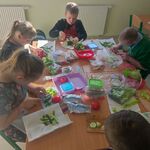 Uczniowie siedzą wokół stołu i przygotowują kanapki z produktów leżących przed nimi na stole.
