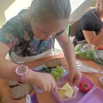 Uczennica siedzi przy stoliku i bierze masło nożem z pojemnika.  Przed nią na stole leżą produkty do przygotowania kanapek.