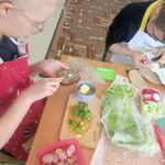 Uczennica siedzi przy stoliku i  smaruje chleb masłem. obok na stole leżą produkty do przygotowania kanapek.