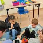 Uczniowie siedzą na dywanie po turecku  w kółku. Z tyłu stoi stół na którym stoi pudełko z układanką.