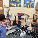 Grupa uczniów siedzi na dywanie i naśladuje nauczycielkę pokazującą jak należy zasłaniać buzię w trakcie kichania,