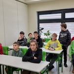 Grupa uczniów  siedzi w ławkach przy stolikach i oglądają prezentację.