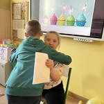 Dziewczynka siedzi na krześle z laurką, chłopiec ją przytula.