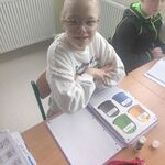 Uczennica siedzi przy stole. Przed nią leży duży zeszyt z wklejoną kartą pracy z pojemnikami na śmieci.