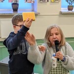 Nauczycielka mówi do mikrofonu. Obok stoi uczeń z gąbką w ręku.