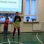 Uczniowie siedzący na ławkach obserwują nauczycielkę i ucznia stojących na tle ekranu z napisem - Obowiązki domowe.