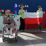 Uczennica na wózku śpiewa do mikrofonu. Z tyłu stoją uczniowie z flagą.