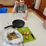 Uczeń siedzi przy stole. Przed nim na stole leży worek z ziemniakami oraz zielona deska na której leżą ziemniaki i obierak. Obok stoi patelnia.