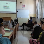 Grupa uczniów siedzi na fotelach w pracowni komputerowej o ogląda webinar wyświetlany na ekranie.