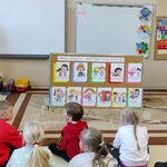 Uczniowie siedzą na podłodze i słuchają pani pedagog. Przed uczniami stoi tablica z kolorowymi obrazkami.