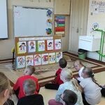 Uczniowie siedzą na podłodze i słuchają pani pedagog. Przed uczniami stoi tablica z kolorowymi obrazkami.