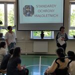 Uczniowie siedzą na ławkach i słuchają pań pedagoga i psychologa stojących na tle wyświetlanego slajdu na ekranie.