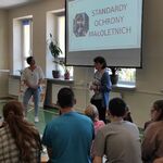 Uczniowie siedzą na ławkach i słuchają pań pedagoga i psychologa stojących na tle wyświetlanego slajdu na ekranie.