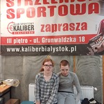 Uczeń i uczennica siedzą  pod dużym napisem Strzelnica sportowa zaprasza.