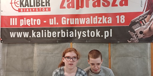 Uczeń i uczennica siedzą  pod dużym napisem Strzelnica sportowa zaprasza.