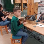 Uczeń siedzi przy stanowisku strzelniczym, w ręku, w ręku trzyma karabinek