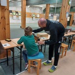 Uczeń siedzi na stanowisku strzelniczym, instruktor pokazuje uczniowi, jak korzystać z broni