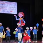 Uczniowie w niebieskich koszulkach stoją na scenie z balonami, dziewczynka ubrana na czerwono na wózku śpiewa do mikrofonu