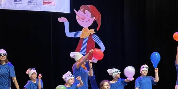 Uczniowie w niebieskich koszulkach stoją na scenie z balonami, dziewczynka ubrana na czerwono na wózku śpiewa do mikrofonu
