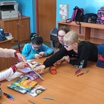 Uczniowie siedzą wokół stołu, autorka prezentuje książkę, uczennica wskazuje ilustrację.