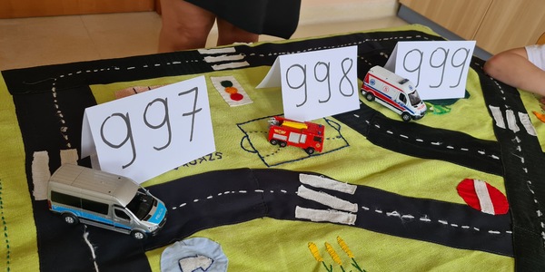 Na makiecie stoją samochody i kartki z numerami alarmowymi.