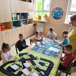 Nauczycielka stoi przy uczniach. Uczniowie siedzą przy stoliku.