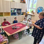 Nauczycielka czyta, uczniowie siedzą przy stole, na stole stoją pojemniki z herbatą.