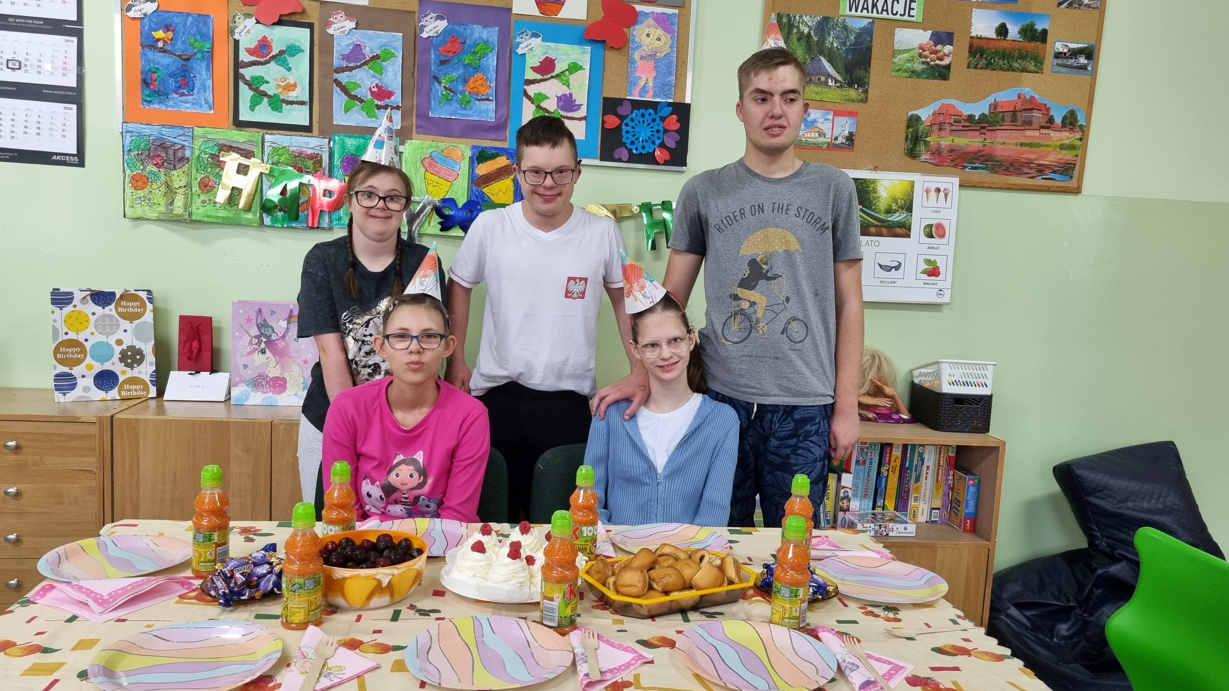 Jubilatki siedzą za urodzinowym stołem. Z tyłu stoją koledzy i koleżanka z klasy.
