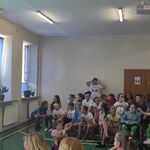 Uczniowie siedzą na ławeczkach i słuchają nauczycielki omawiającej  slojd wyświetlany na ekranie.