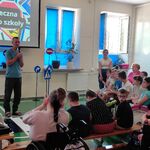 Uczniowie siedzą na ławeczkach i słuchają nauczyciela mówiącego do mikrofonu.