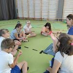 Grupa uczniów i nauczycielka siedzą w kole na podłodze. Uczniowie wykonują prezentowane przez nauczycielkę ćwiczenie.