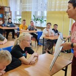 Grupa uczniów siedzi w ławkach. Nauczycielka  pokazuje obrazki i mówi do uczniów.
