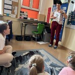 Nauczyciel stoi z kartką w ręku stoi przed uczniami i wskazuje na znak zapytania widniejący na papierowej  planszy.
