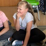 Troje uczniów siedzi na dywanie. Jeden z nich uśmiecha się.
