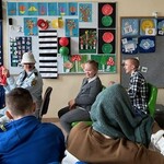 Nauczyciel przebrany za kierowcę siedzi na krześle. Za nauczycielem siedzą uczniowie.