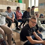 Uczniowie siedzą na dywanie i na krzesłach.