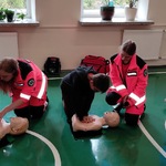 Uczniowie siedzą na ławkach i obserwują kolegów wykonujących masaż serca pod kontrolą ratowników medycznych.