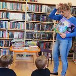 Na tle regałów bibliotecznych stoi pani bibliotekarka. Na podłodze siedzą uczniowie.