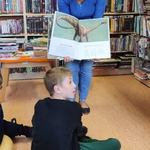 Pani bibliotekarka pokazuje uczniowi otwartą książkę ze zdjęciem dinozaura.