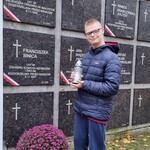 Uczeń trzyma znicz w rękach na tle tablic nagrobkowych.