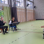 Uczniowie siedzą na ławkach i oglądają występ muzyczny.