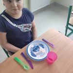 Uczennica siedzi przy stole na którym stoi talerz, kubek i leżą sztućce.