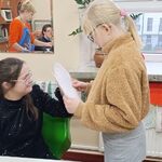 Uczennica przebija szpilką talerzyk papierowy z wróżbą trzymany przez koleżankę.