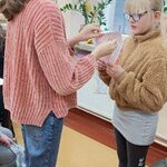 Uczennica przebija szpilką talerzyk papierowy z wróżbą trzymany przez koleżankę.