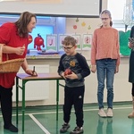 Nauczycielka stoi z koszykiem, obok uczeń z jabłkiem. W tle nauczycielka z uczniem.