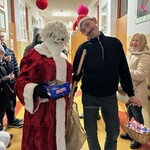 Mikołaj stoi i trzyma Pudełko z batonikami obok stoi „Śnieżynka” w czapce mikołajowej, trzyma w ręku kosz z cukierkami.