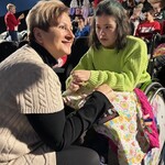 Na pierwszym planie nauczycielka uśmiecha się i pochyla się do uczennicy siedzącej na wózku.