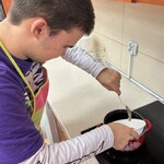 Uczeń łyżką zgarnia z miski miód do garnka.