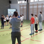 Grupa uczniów na sali gimnastycznej wykonuje ćwiczenia prezentowane na ekranie.