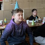 Uczniowie w kolorowych czapeczkach siedzą przy urodzinowym stole.